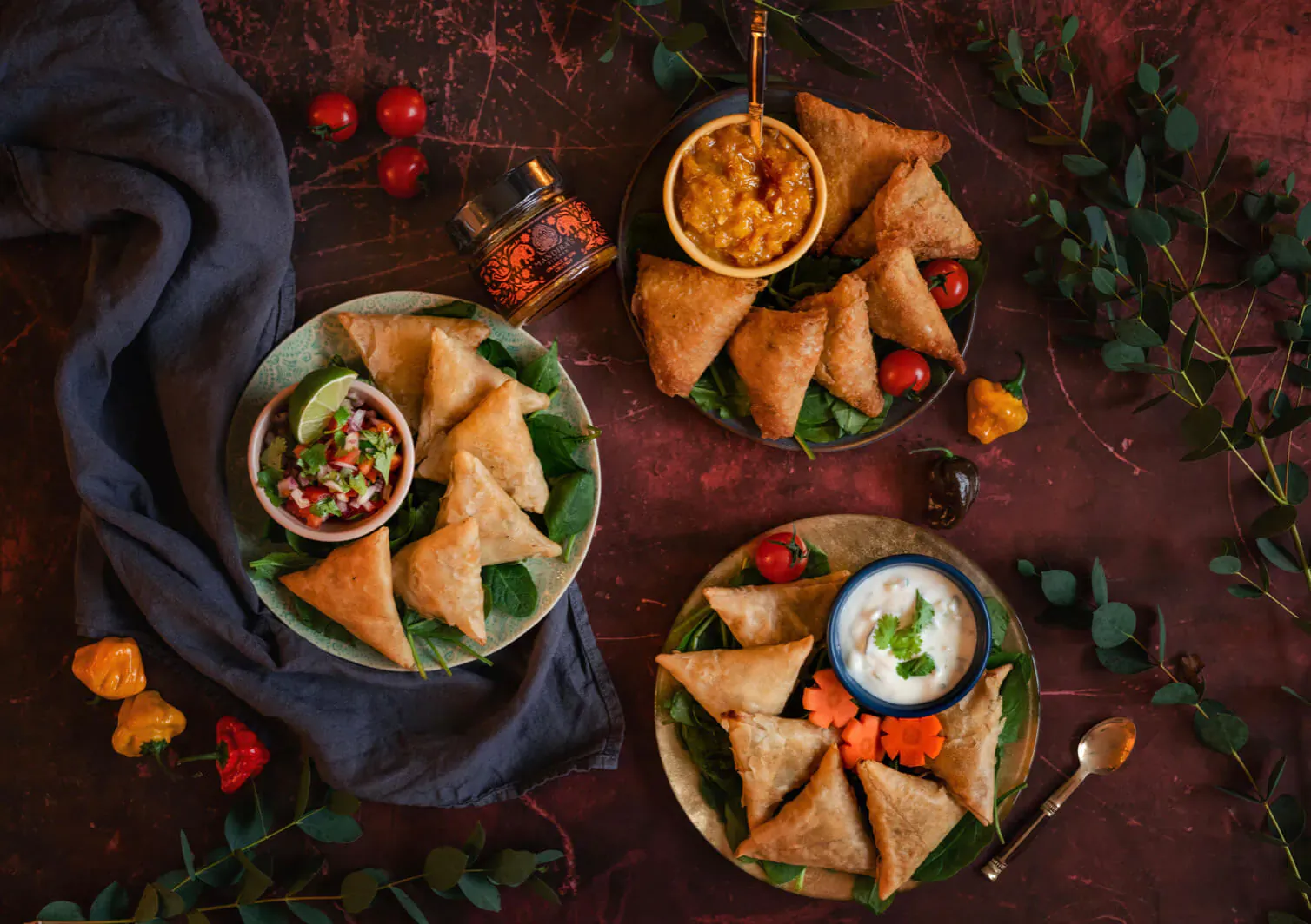 Assortment of golden samosas on plates with chutneys, yogurt dip, garnished with herbs and peppers on dark textured surface