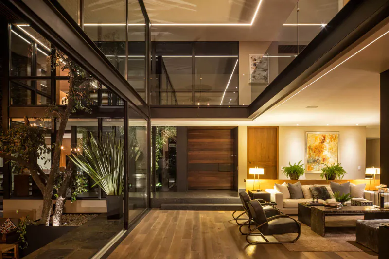 Modern Mexican home interior with glass walls, courtyard tree, wooden door, warm-lit leather seating area