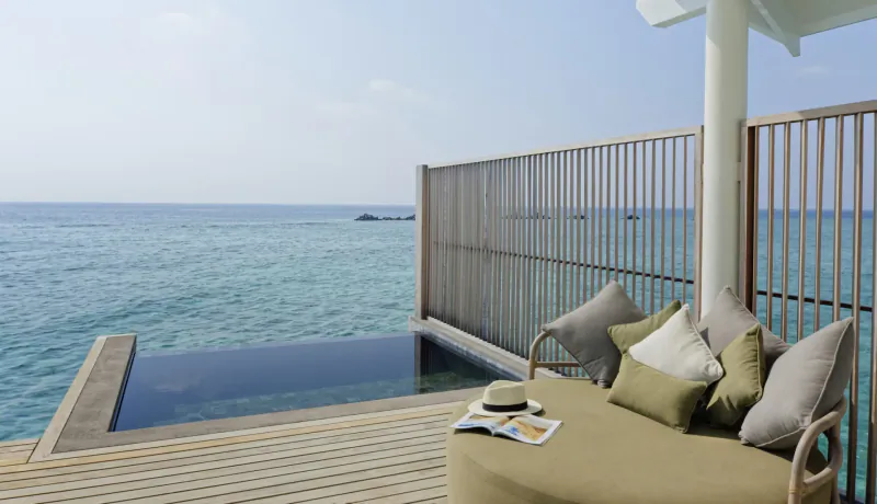 Luxurious overwater villa deck in Maldives with infinity pool, turquoise ocean view, and cushioned lounge seat with open magazine.