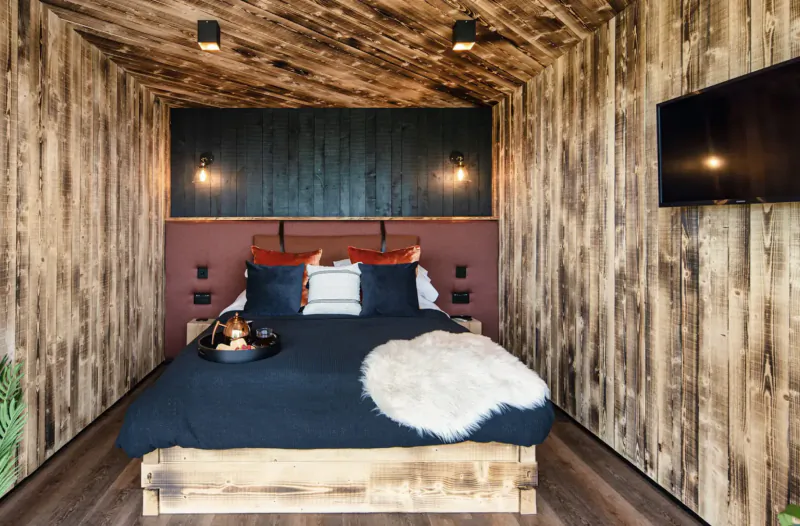 Cozy wooden interior of luxury glamping pod with king bed, fur throw, pillows, tray, and wall-mounted TV.