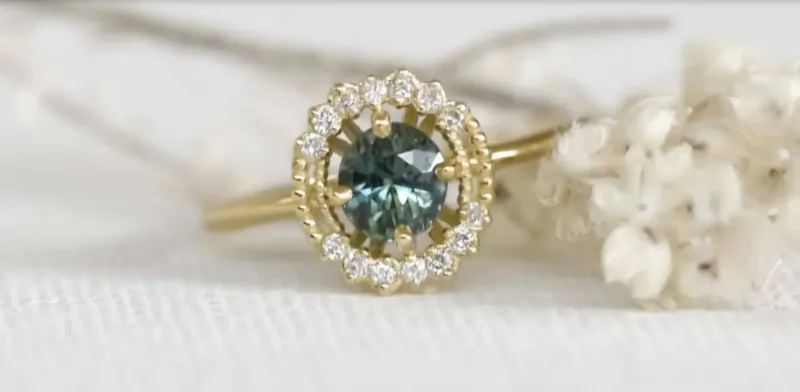 Close-up of gold ring with teal center gem and diamond halo, on white surface with white dried flowers