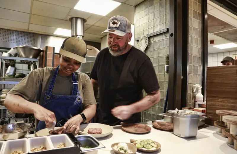 Black woman in apron chopping burger patty and bearded man in cap preparing food on counter in kitchen