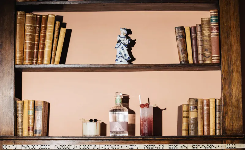 Wooden bookshelf in Home House club with vintage books, blue porcelain figurine, gin bottle, red cocktail, and white candle.