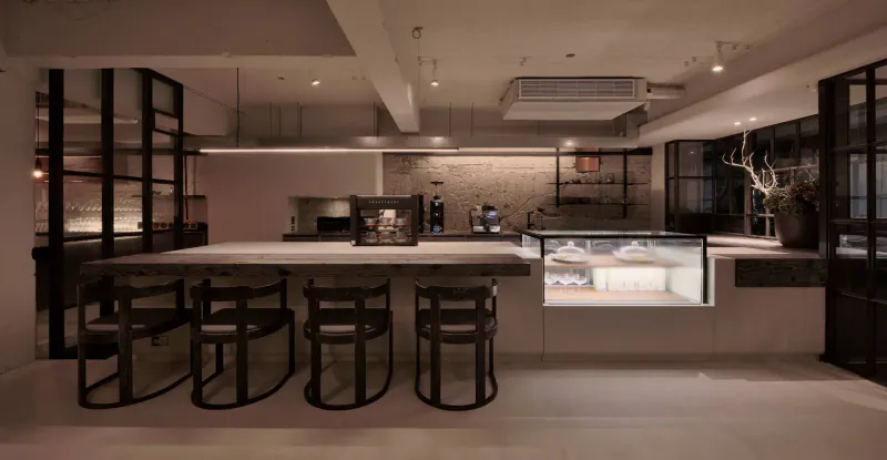 Stylish contemporary restaurant interior with long black metal bar table, stools, display case, and plants.