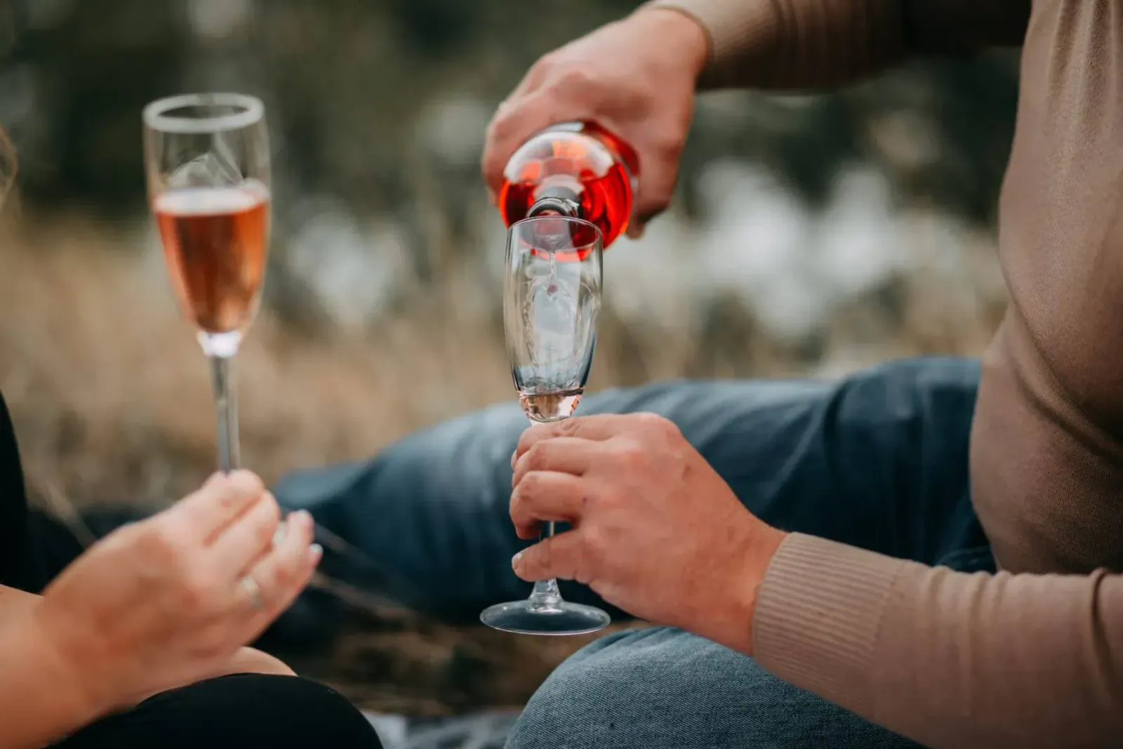 Couple at posh outdoor picnic toasting with champagne flutes, man pouring rosé into woman's glass amid grass.