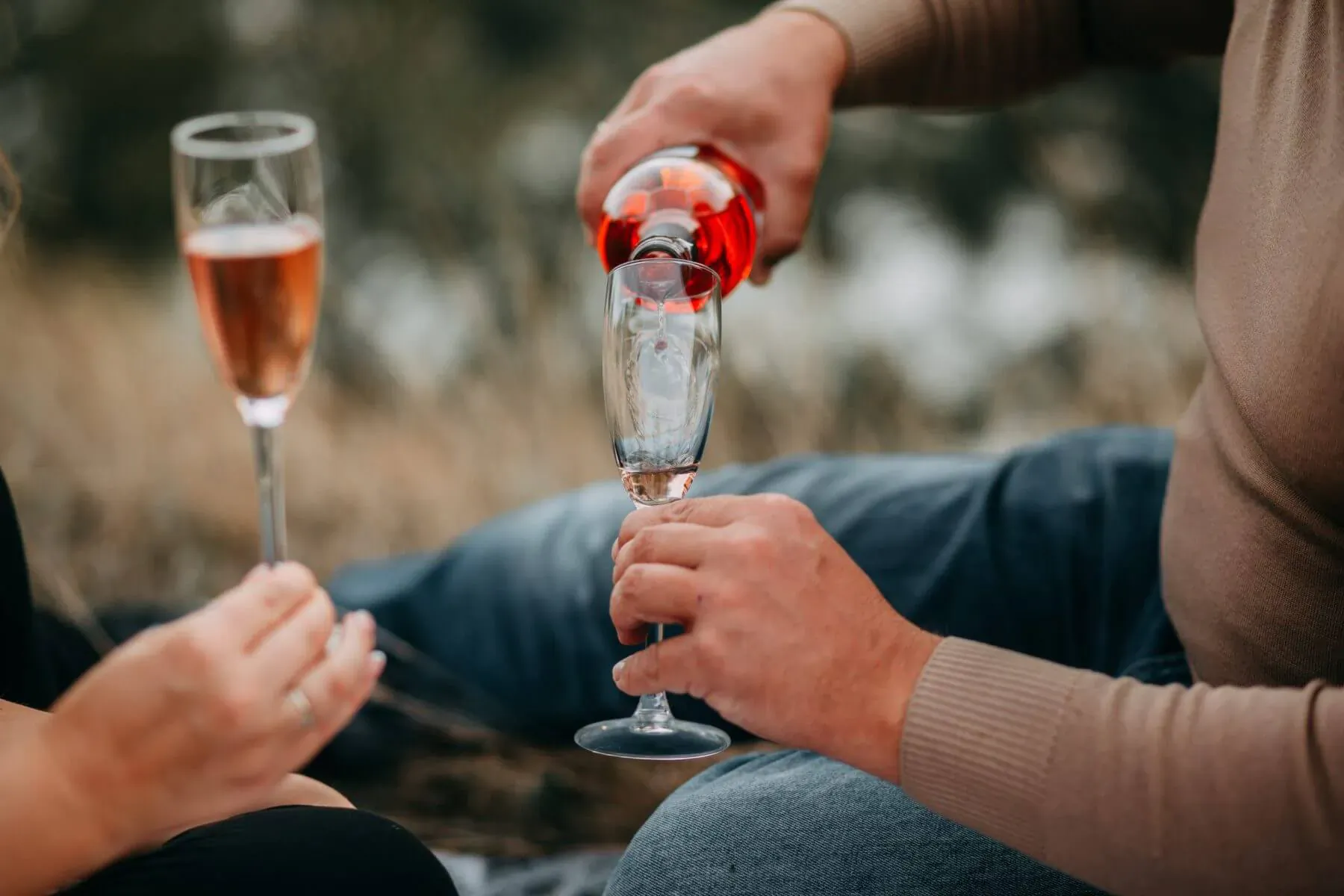 Couple at posh outdoor picnic toasting with champagne flutes, man pouring rosé into woman's glass amid grass.