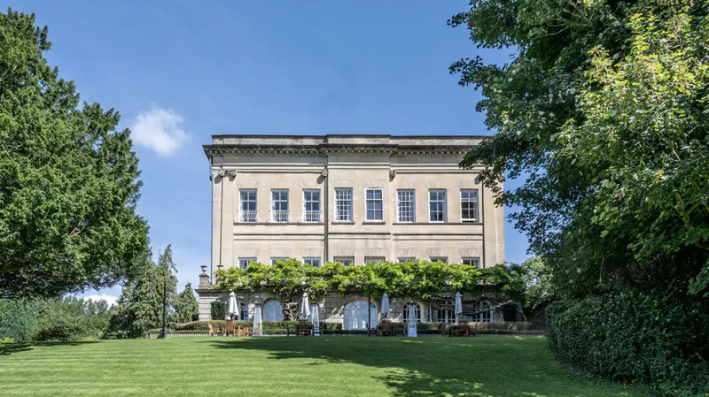Bailbrook House Hotel, grand pale stone country mansion in Bath UK, with wisteria terrace, umbrellas, and lush green gardens under blue sky.