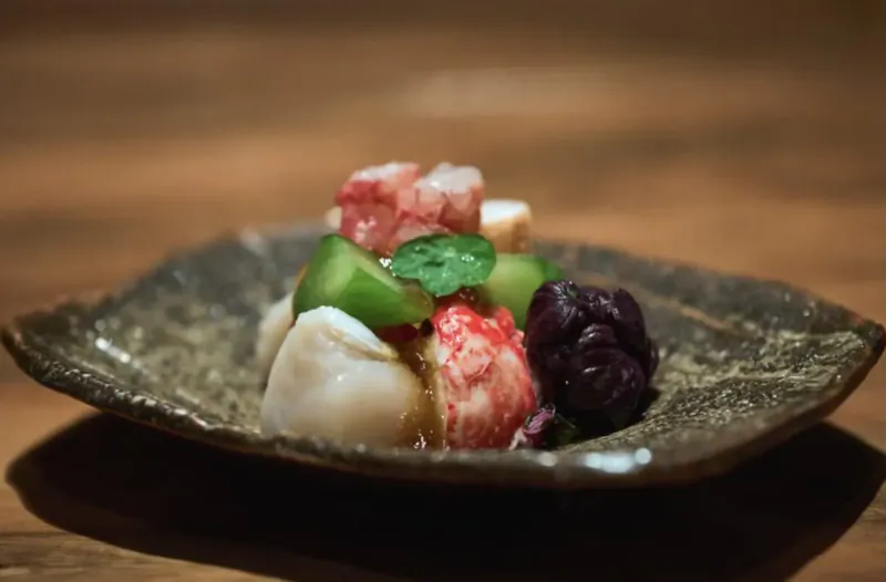 Japanese dish on square plate: crab, white seafood, green melon, plums, sauce on wood table