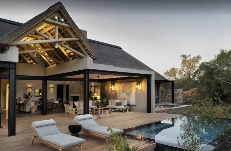 Monwana Game Lodge's modern thatched eco-lodge exterior with outdoor lounge, pool, and savanna surroundings at dusk.