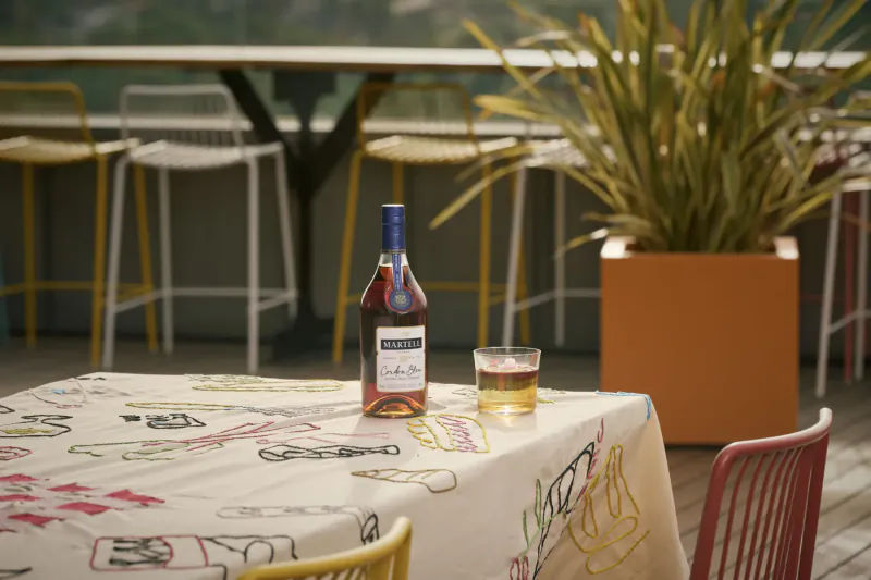 Martell cognac bottle and whiskey glass on colorful tablecloth at L’Indigo bar balcony with yellow chairs and plants