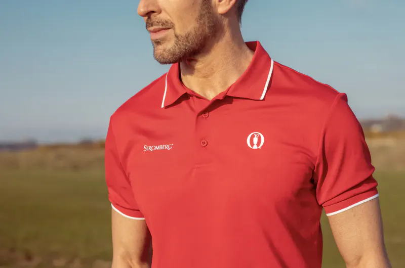Man in red 'Synergy' polo shirt with white trim and logo, posing outdoors on green field