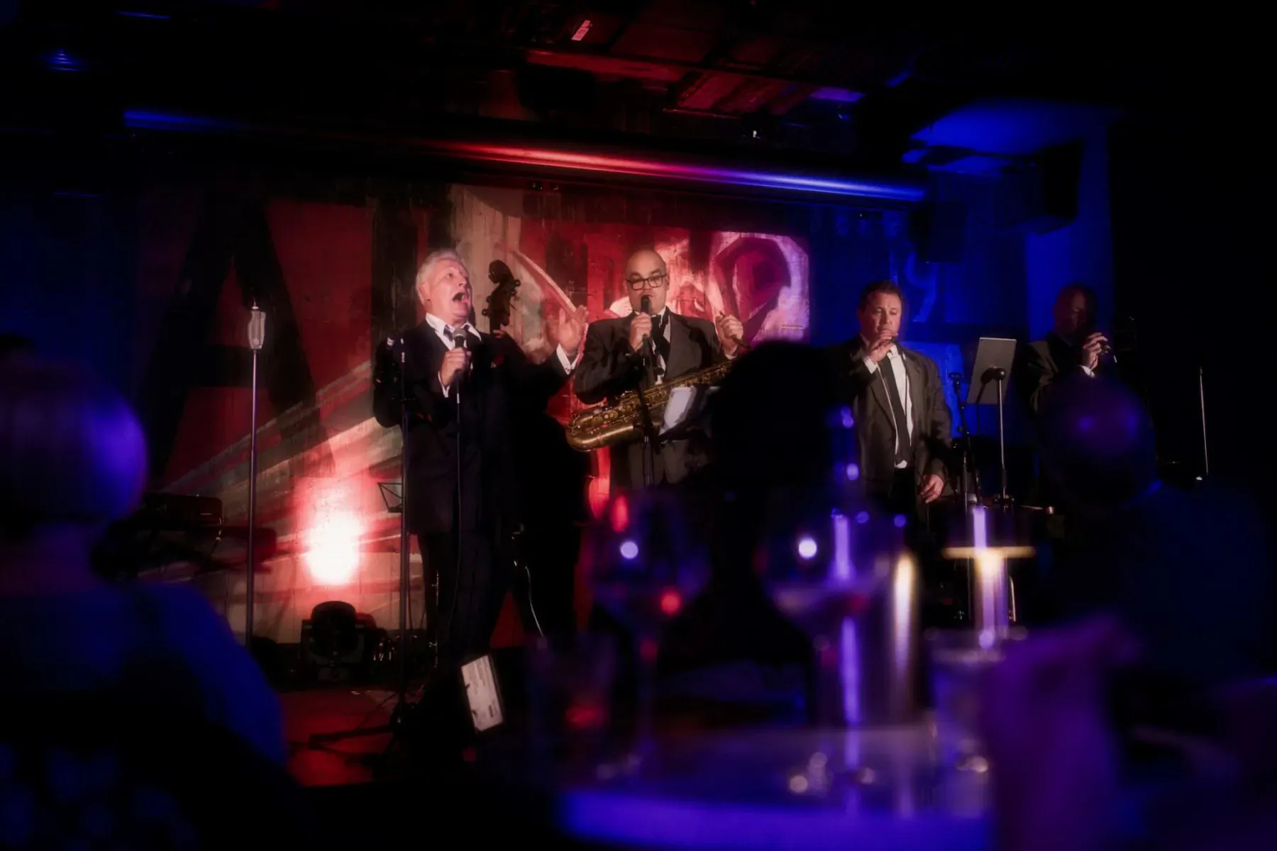 Band performing live on stage at luxury soiree: singer at mic, saxophonist, suited musicians, red lighting.