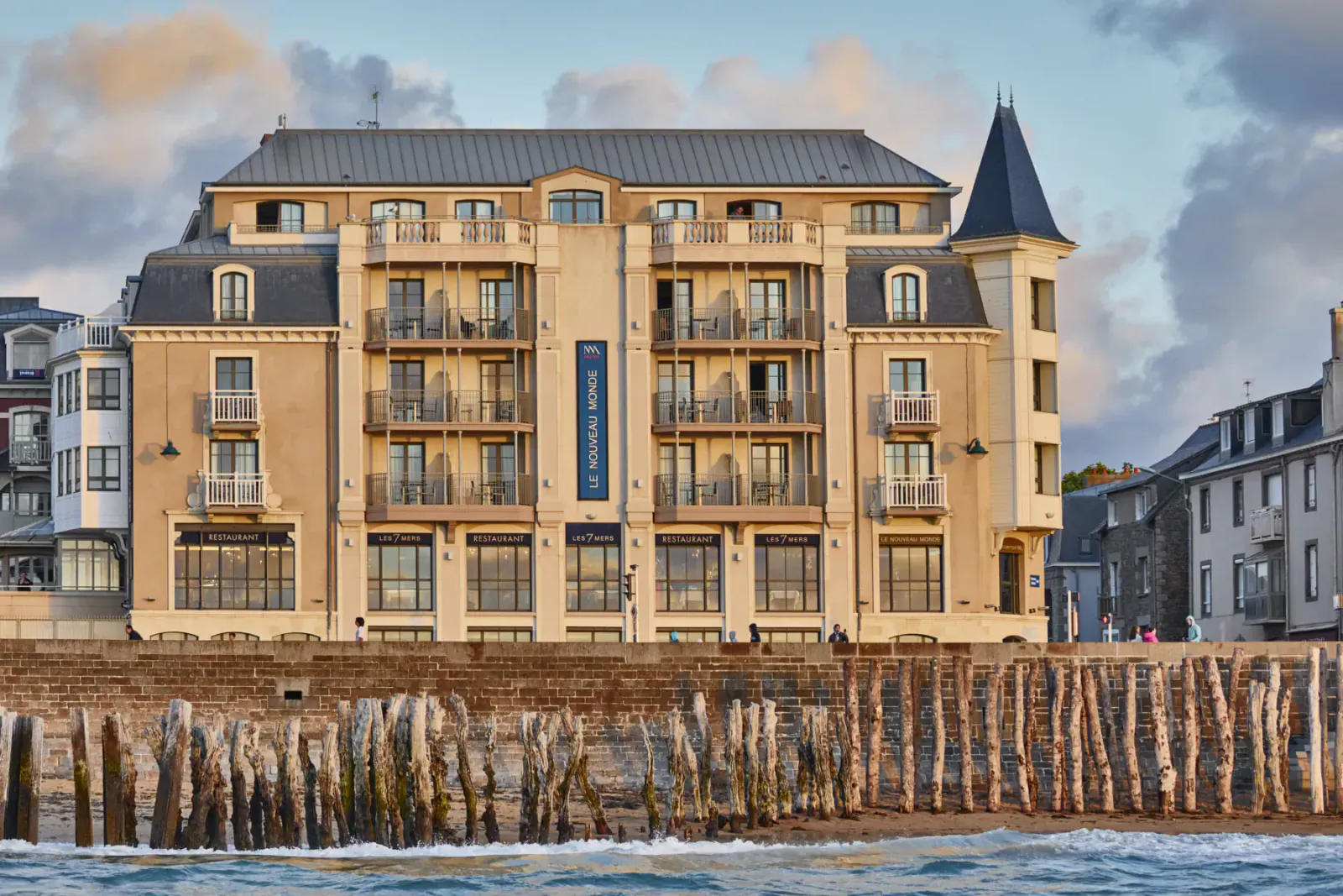 Hotel Nouveau Monde Saint-Malo, a beige multi-story seafront hotel with balconies and tower, at sunset by the ocean.
