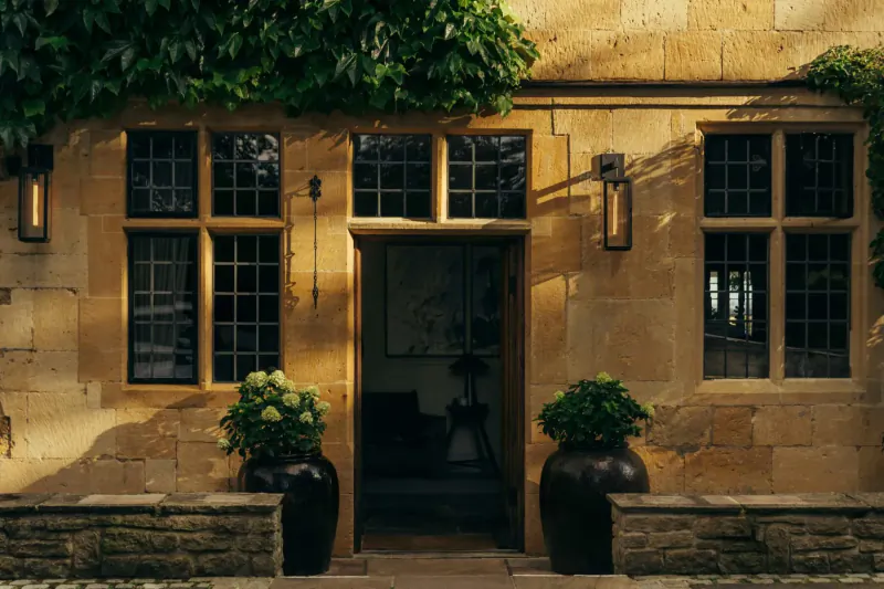 Open entrance to a luxurious stone cottage with ivy-covered walls, large windows, lanterns, and potted plants.