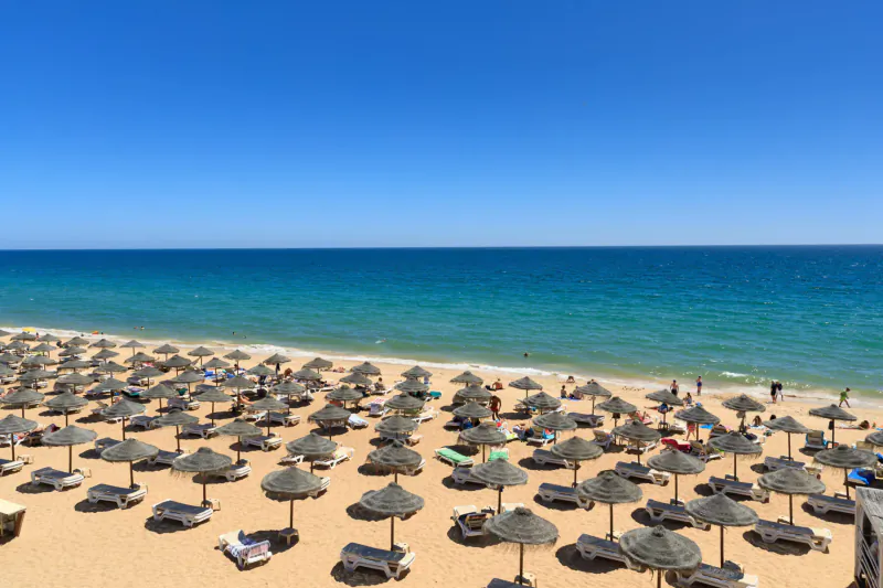 Aerial view of Vale do Lobo beach resort in Portugal with rows of colorful umbrellas on golden sand and turquoise sea under blue sky.