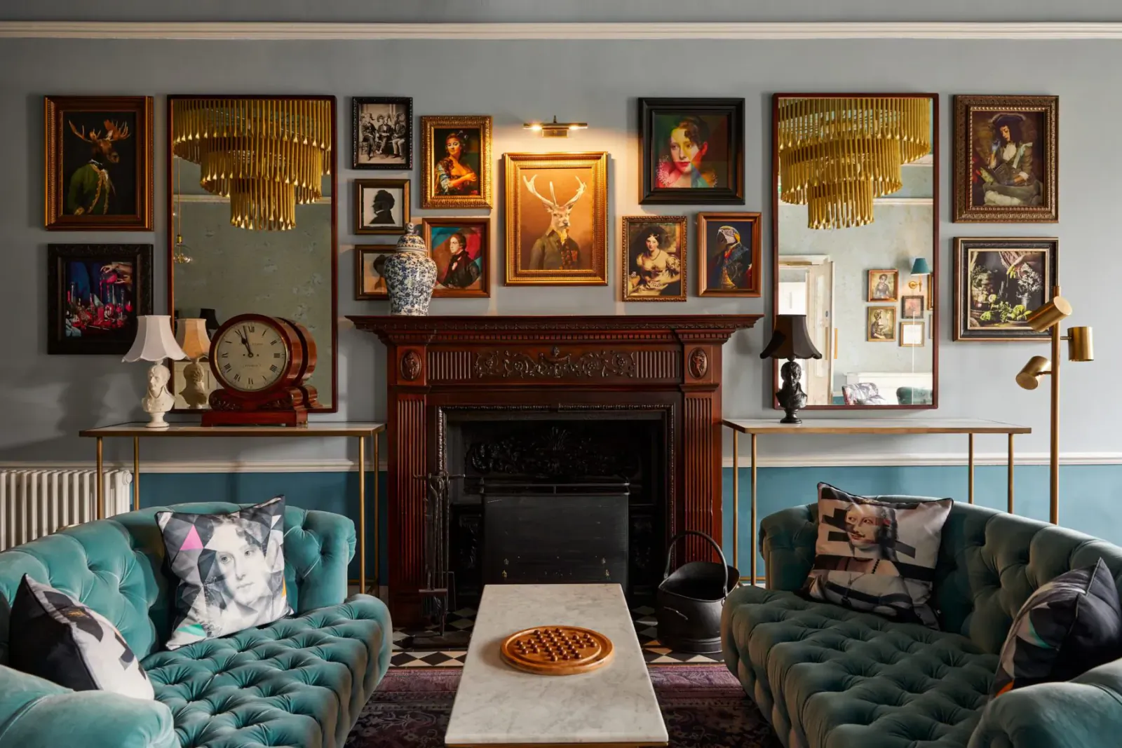 Elegant Hotel Indigo Bath lounge with teal velvet sofas, ornate wooden fireplace, eclectic art on walls, and brass lamps.
