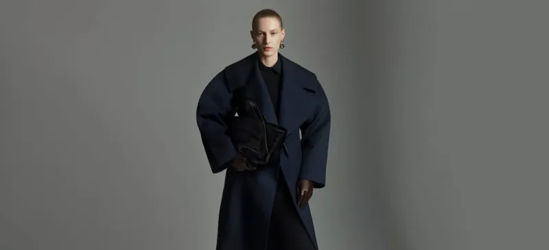 Model poses confidently in oversized navy Scanlan Theodore coat holding black bag, gray background