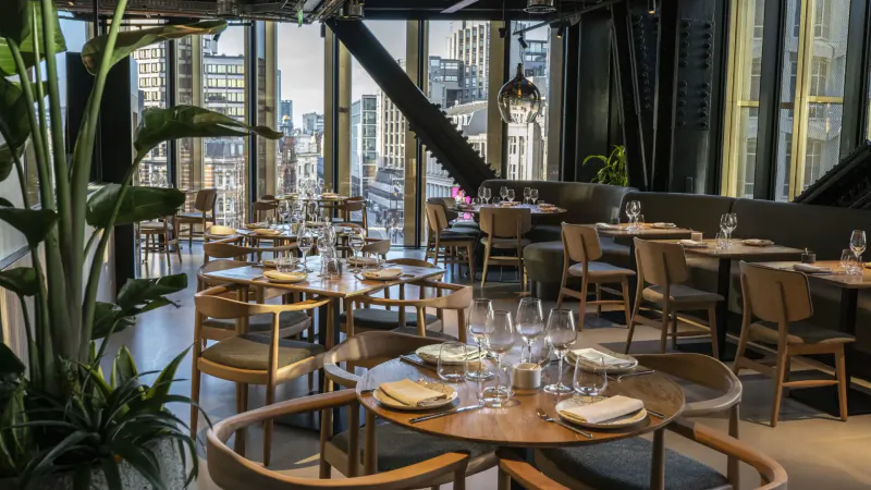 Cavo rooftop Mediterranean restaurant in Central London: elegant wooden tables set with glassware, plants, city skyline view.