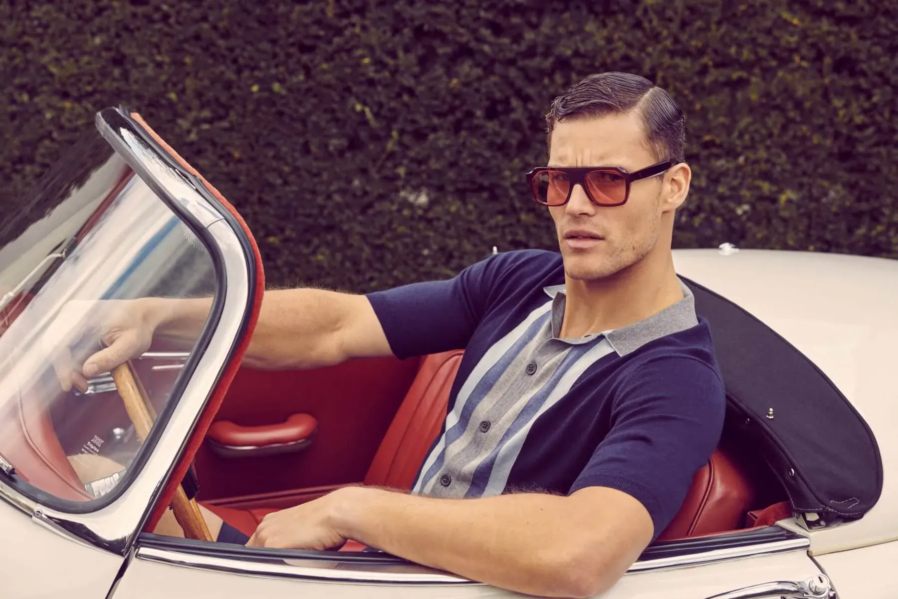 Stylish man with orange sunglasses driving white convertible sports car, hedgerow backdrop