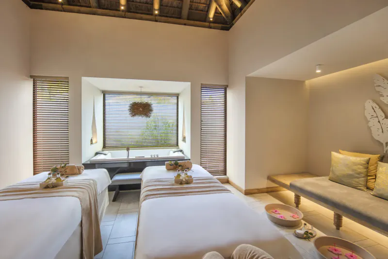 Luxurious spa treatment room at Anantara Iko Mauritius Resort with two massage beds draped in beige linens, flower bowls, and tropical decor.
