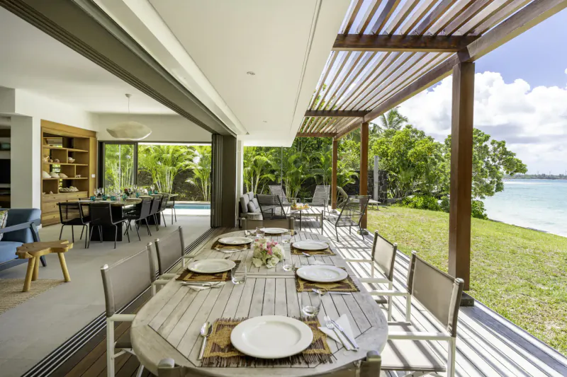 Luxury villa's open-plan dining area with long wooden table set for meal, overlooking turquoise sea and tropical garden in Mauritius.
