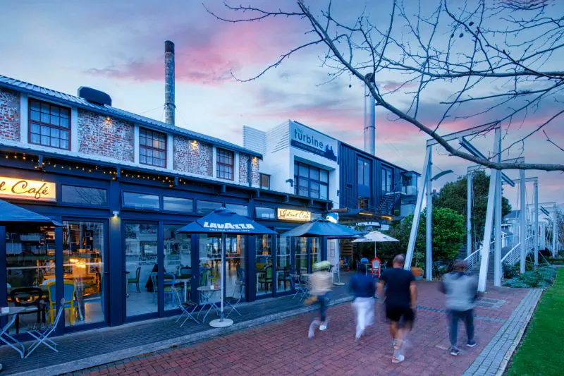 Turbine Boutique Hotel & Café on Thesen Islands, Knysna: blue umbrellas, patrons walking on red brick path at sunset with smokestack.