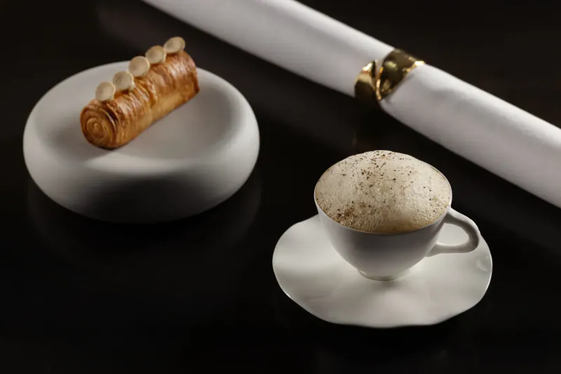 Pastry roll on white plate with nuts beside cappuccino with foam on saucer and napkin with gold ring