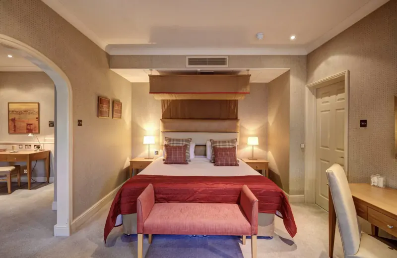 Luxurious hotel bedroom with red king bed, plaid pillows, bench, desk, and arched doorway