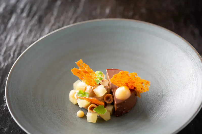 Close-up of chocolate mousse cake with diced apples, hazelnuts, and crispy orange tuiles on a blue plate.