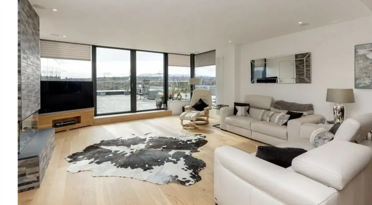 Modern penthouse living room with white leather sofas, cowhide rug, fireplace, TV, and panoramic city views through large windows.