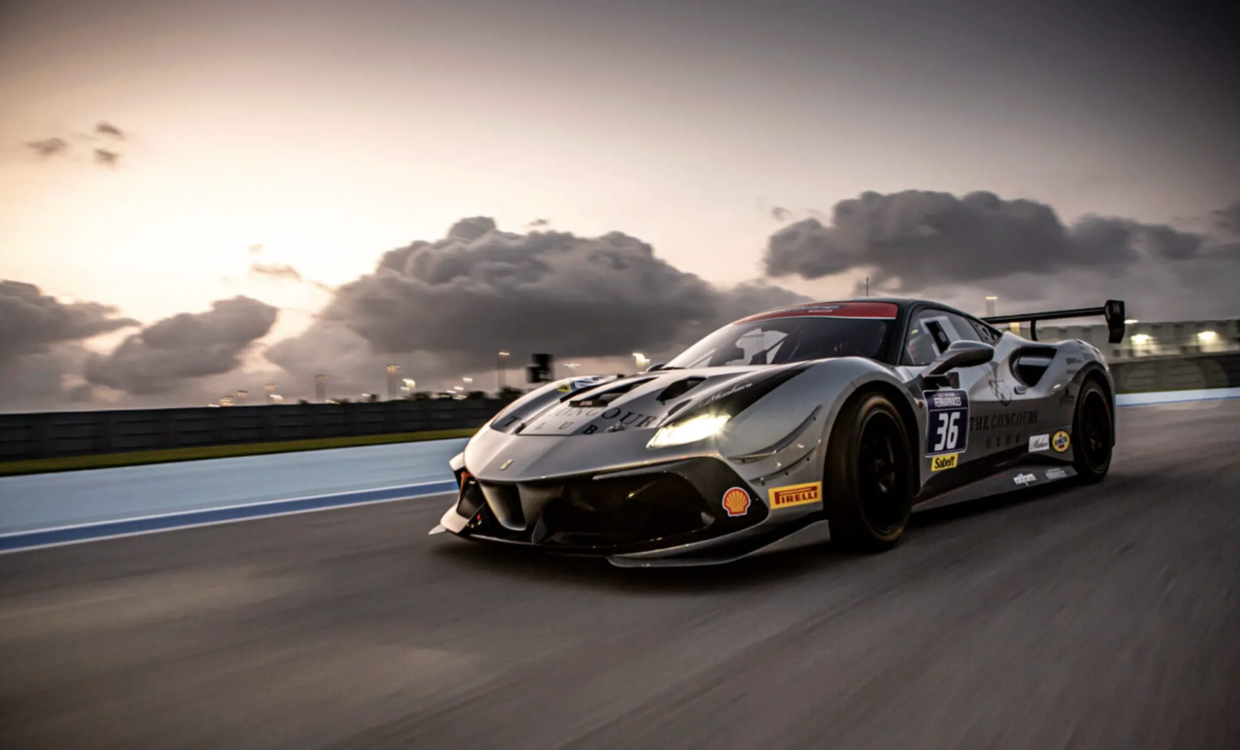 Silver Ferrari 488 GT3 race car speeding on track at sunset with dramatic clouds, number 63 on side.