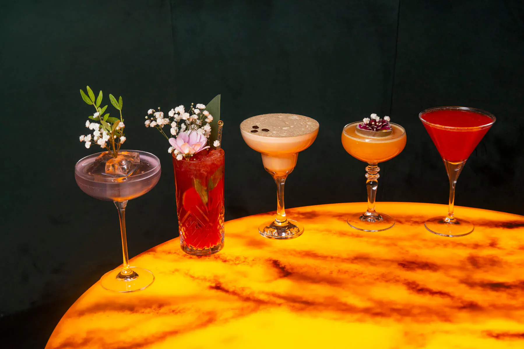 Four colorful alcohol-free cocktails garnished with flowers and herbs on a glowing orange table at Coco Grill & Lounge.