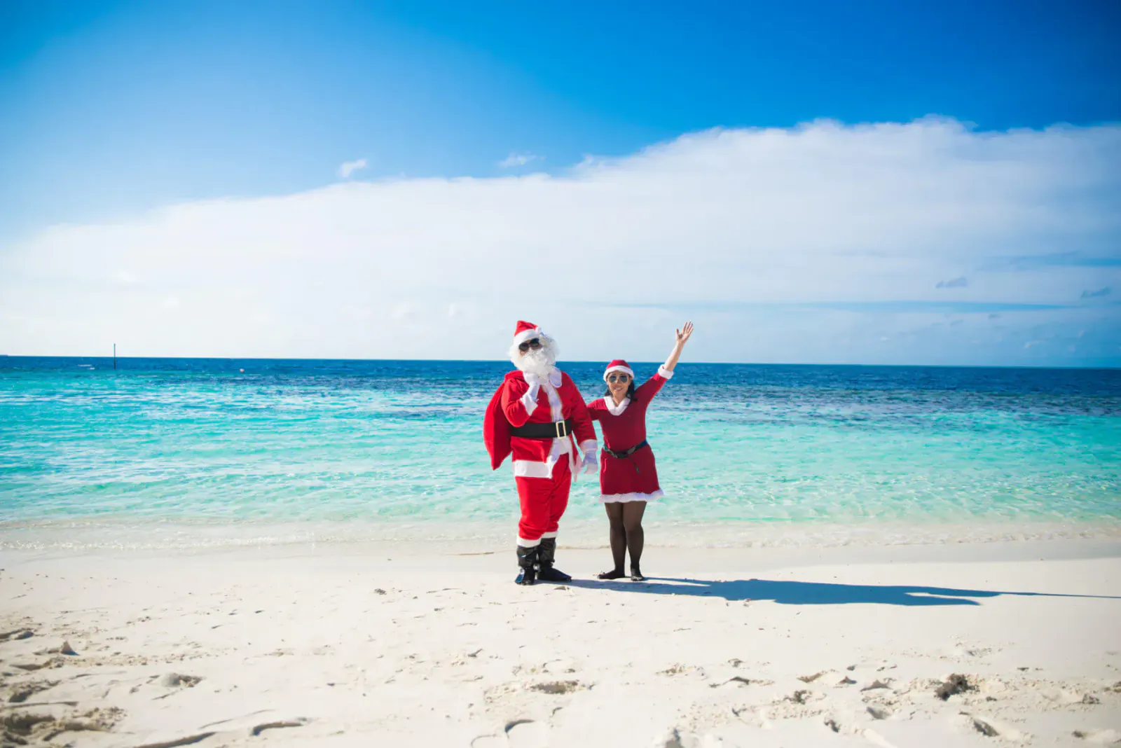 Santa and girl in red dress waving on tropical beach with turquoise sea and blue sky