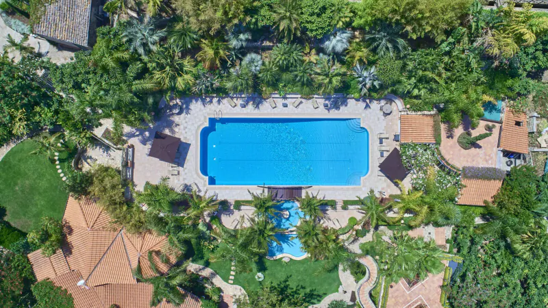 Aerial view of Wish Sicily luxury villa with rectangular blue pool, lush tropical gardens, and terracotta buildings.