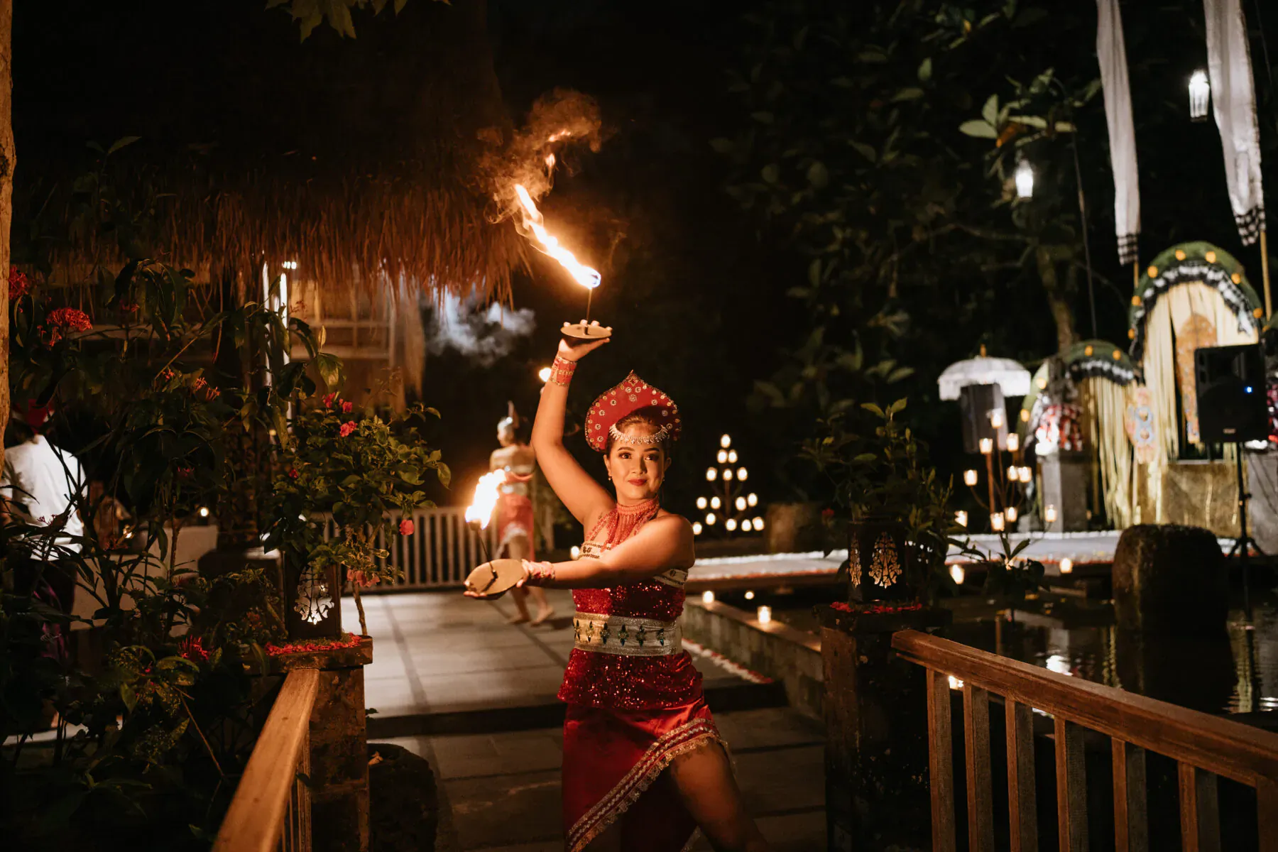 Balinese dancer in red costume performs fire poi on wooden bridge at Kepitu Restaurant, night with lanterns and tropical plants