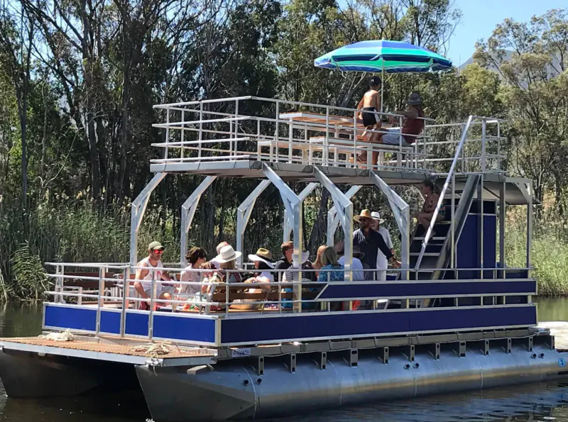 Pontoon boat with people relaxing on decks under green-blue umbrella, cruising tree-lined Klein River.
