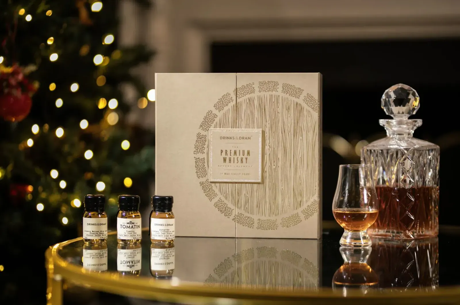Premium Whisky Advent Calendar box with four sample bottles, crystal decanter, and whisky glass on table amid Christmas lights and tree.
