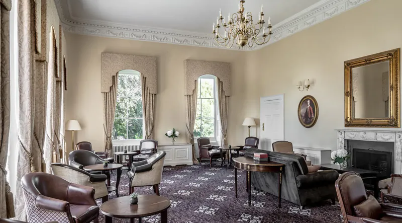 Elegant drawing room at Bailbrook House Hotel with leather armchairs, tables, chandelier, large windows, and fireplace.