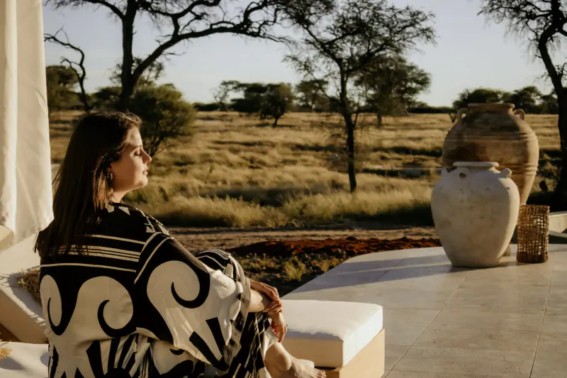 Woman in black and white patterned dress sits on patio at Namibian savanna luxury villa, gazing at acacia trees and large pottery vase at sunset.