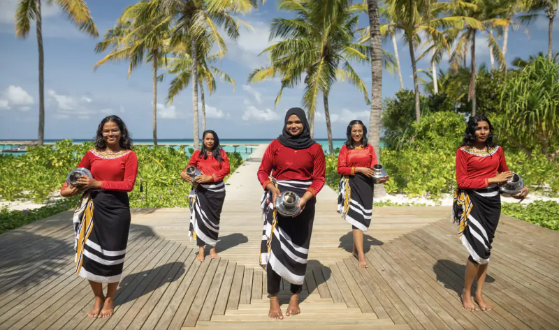 Five women in red tops, black-white skirts, and hijabs hold drums on a wooden path amid Maldives palm trees and beach.
