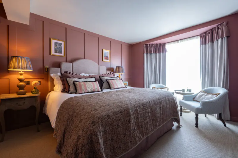 Elegant blush pink bedroom with king bed, fringed lamps, armchairs, and large window drapes.
