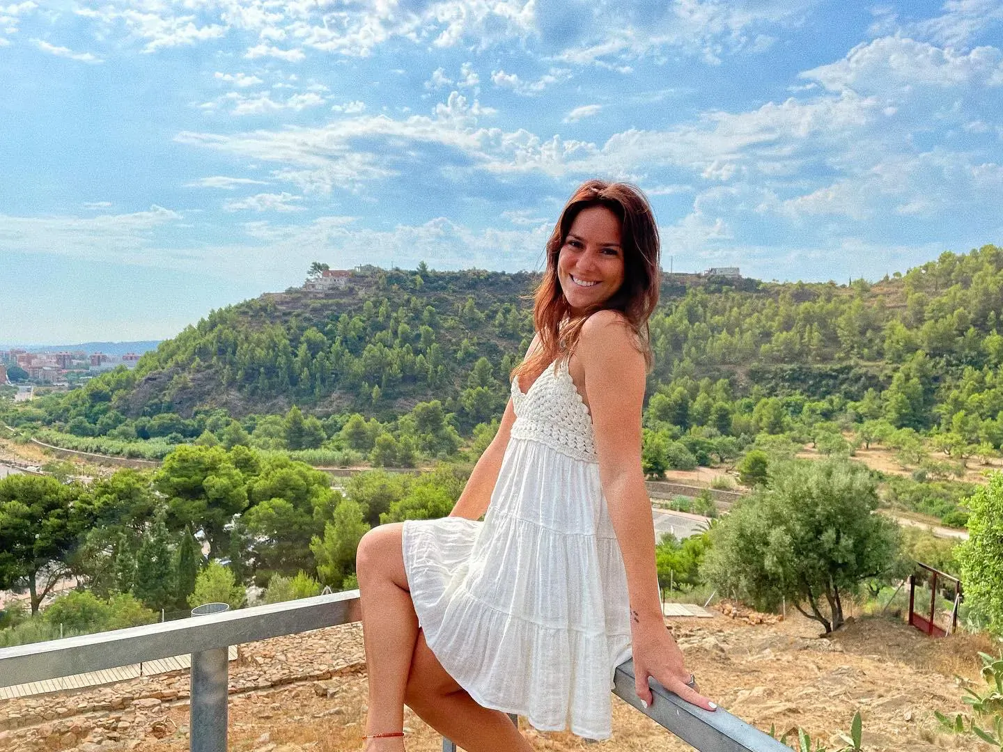 Dayami Rivera in white tiered sundress, smiling and leaning on railing with hills and blue sky behind her
