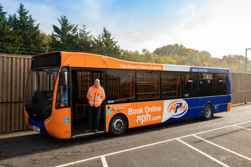 Orange Airport Parking Gatwick bus with hi-vis worker standing at open door, 'Book Online apn.com' on side, at Gatwick Airport.