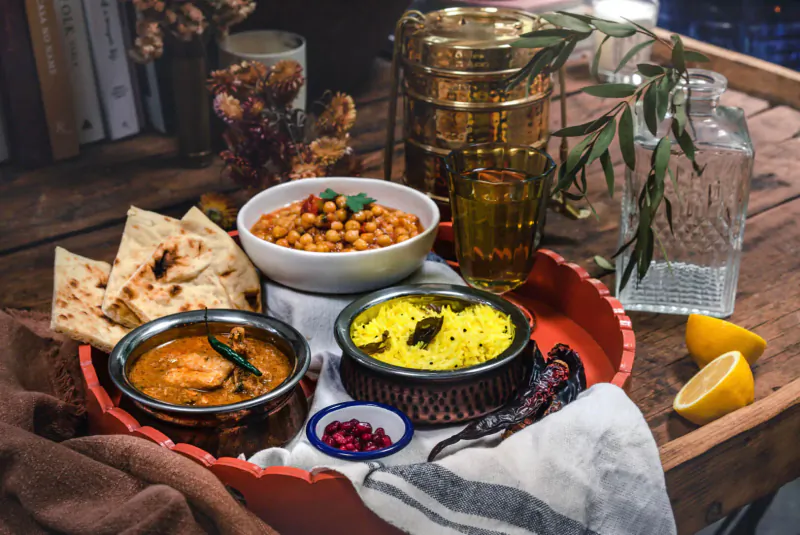 Indian thali on wooden table: chickpea curry, yellow dal, chicken curry, naan, served in brass bowls with lemon wedges