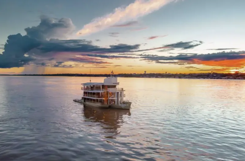 Three-story paddlewheel boat on river at sunset with dramatic clouds and orange sky.