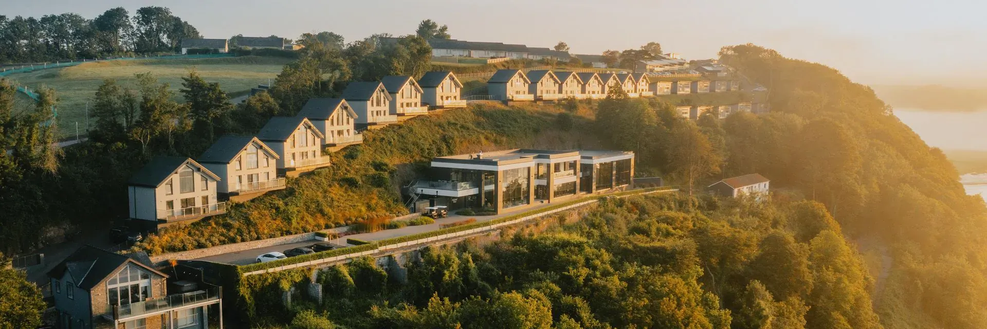 Aerial view of Dylan Coastal Resort's white chalet-style villas on green cliffside overlooking river at golden hour sunset, South Wales.