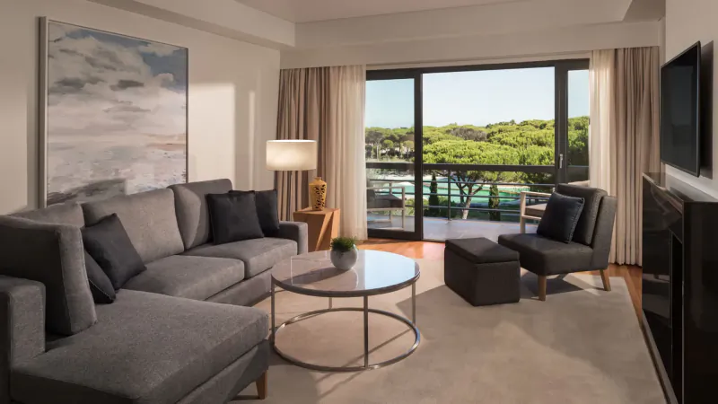Spacious modern hotel living room with gray L-shaped sofa, glass coffee table, lamps, and balcony view of green landscape and water.