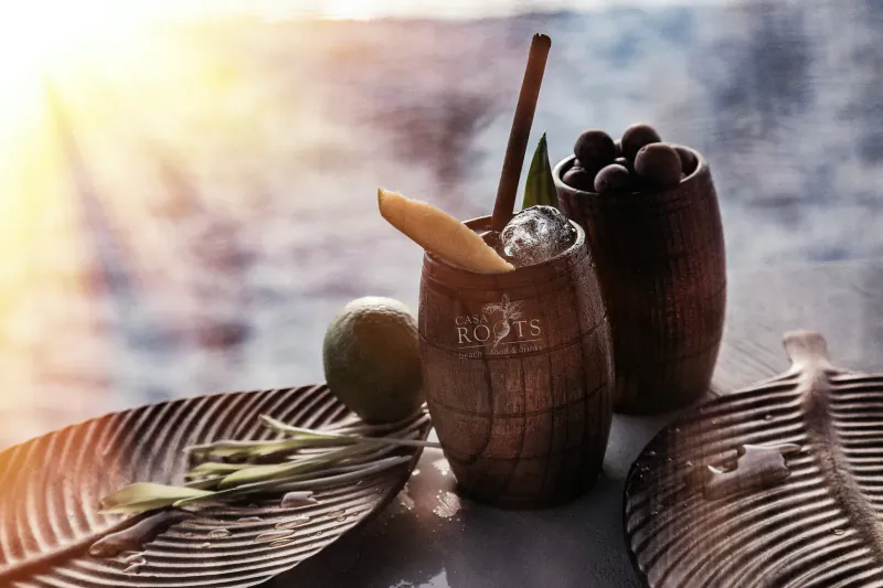 Roots cocktail in wooden barrel glass with lime wheel, ice, bamboo straw, garnished with lime and beans on bamboo plates