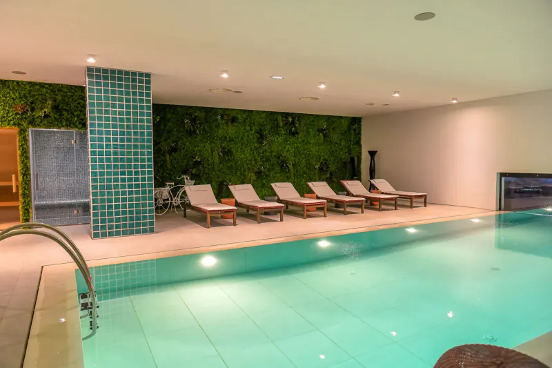 Indoor pool with turquoise tiles, green wall, white lounge chairs, sauna door, and soft lighting