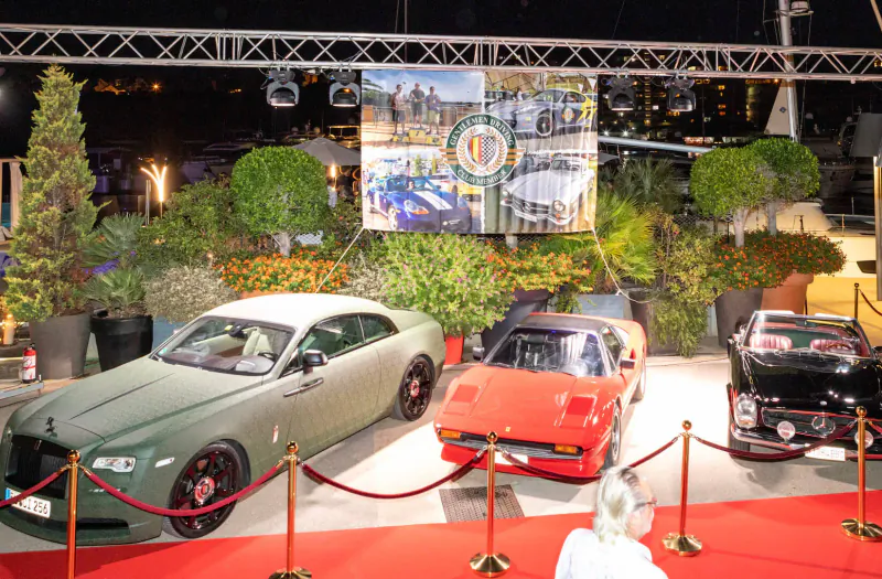 Luxury car exhibition at night: green Rolls-Royce, red Ferrari, black classic, white coupe on red carpet amid plants and banners.